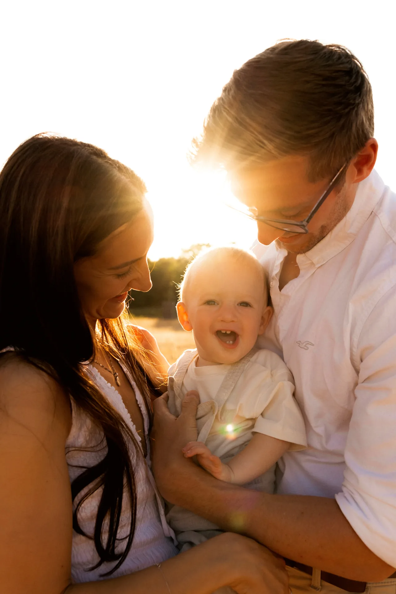 Mother, father and smiling baby dressed in white and cream, backlit by golden-hour sun in an open field - family photography Kingston upon Thames