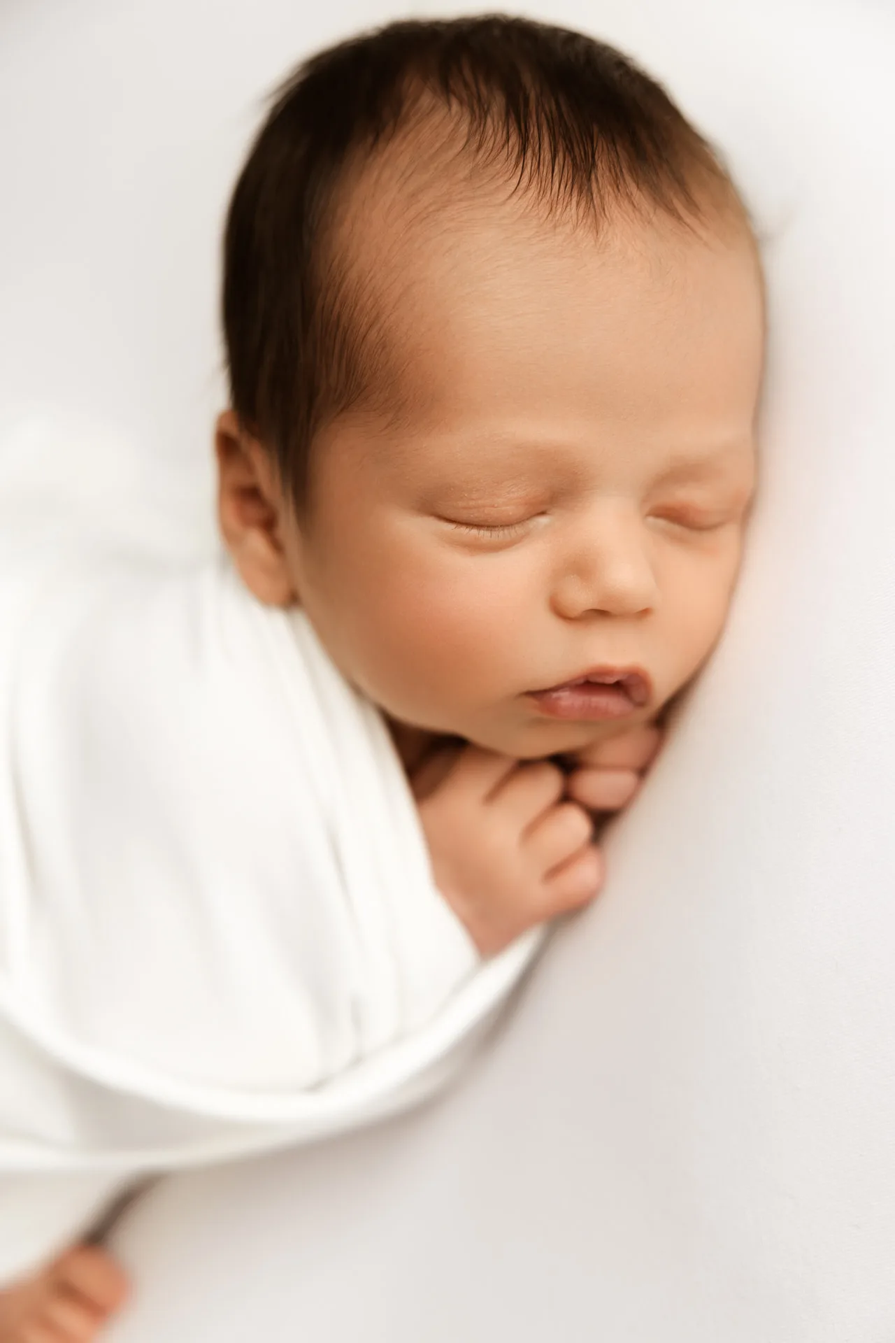 Newborn baby with dark hair sleeping peacefully, swaddled in soft white fabric - newborn photography Kingston upon Thames