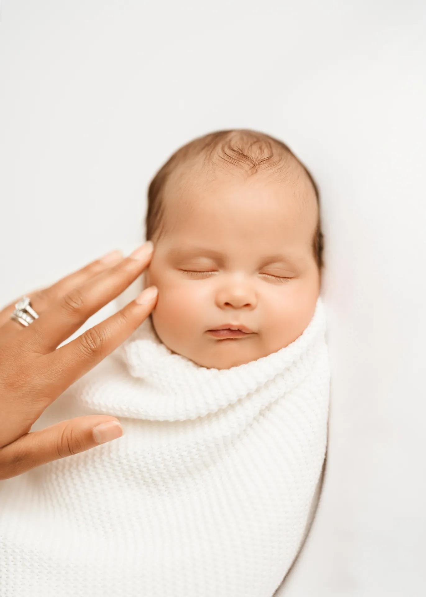 Sleeping newborn baby swaddled in white knit wrap with mother's hand gently touching face - newborn photographer Kingston
